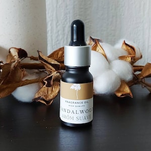 Op de afbeelding: Een klein zwart flesje met een zilveren dop en een druppelaar. Het flesje heeft een bruin etiket met de tekst "FRAGRANCE OIL HIGH QUALITY SANDALWOOD AROM SUAVE".