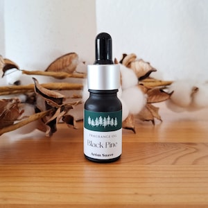 Op de afbeelding: Een klein zwart glazen flesje met een zilveren dop en een druppelaar. Het flesje heeft een groen etiket met de tekst "Fragrance Oil Black Pine Arom Suave" en een klein afbeelding van dennenbomen.
