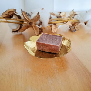 Puede incluir: Una pastilla de jabón cuadrada y marrón descansa sobre un plato dorado en forma de pétalo. El plato está sobre una superficie de madera clara. Tallos de algodón y vainas secas en el fondo, creando una estética natural y de spa.