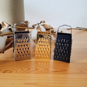 Può includere: Tre piccoli grattugiati per formaggio in metallo, uno argentato, uno dorato e uno nero, su una superficie di legno.