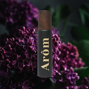 Puede incluir: Una botella negra con una tapa de madera y la palabra "Arom" en letras doradas. La botella está delante de una flor morada.