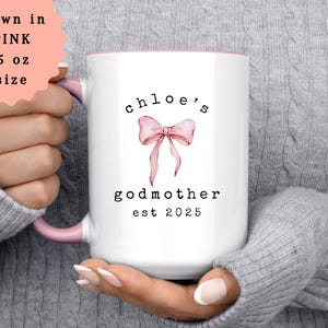 May include: White ceramic mug with a pink interior and handle. The mug features the text "chloe's godmother est 2025" with a pink bow graphic. The mug is 15 oz in size and shown in pink.