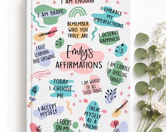 May include: A white hardcover journal titled Emily's Affirmations, featuring colorful abstract shapes, small stars, and botanical line drawings. The cover displays various positive phrases like I Am Enough, I Am Brave, and I Deserve Happiness.