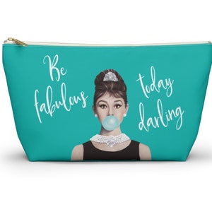 May include: A turquoise zippered pouch with a white graphic of Audrey Hepburn blowing a bubble. The text "Be fabulous today darling" is printed on the pouch.
