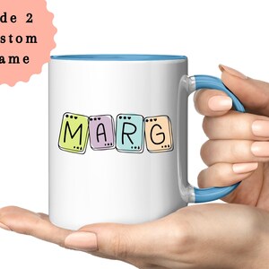 May include: A white ceramic mug with a light blue interior and handle. The mug features the name "MARG" in colorful, square letter tiles. The text "Side 2 Custom Name" is on a pink background in the upper left corner.