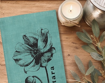 May include: A teal-colored journal with a textured fabric cover featuring a black ink-style illustration of a large poppy flower and a bud. The name "tara" is printed in black lowercase letters vertically next to the flower stem.