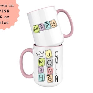 May include: Two white ceramic mugs stacked on top of each other. The top mug has the name "MARG" spelled out in colorful letter tiles. The bottom mug has the words "MOM" and "QUEEN" with a crown graphic. Both mugs have pink handles.