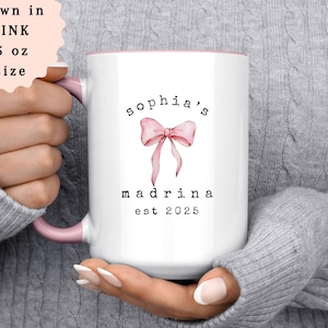 May include: White ceramic mug with a pink interior and handle. The mug features the text "sophia's madrina est 2025" with a pink bow graphic. The mug is 15 oz in size and is shown in pink.