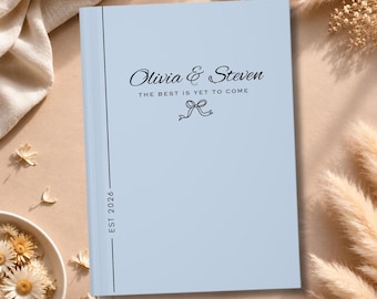 May include: A light blue hardcover book featuring black text that reads Olivia & Steven, The best is yet to come, and EST 2026, accompanied by a small black line drawing of a bow centered below the text and a vertical line along the left side.