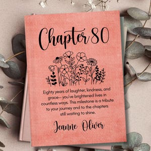 May include: A coral-colored book cover with the words "Chapter 80" in elegant script. Below the title is a floral illustration. The text reads, "Eighty years of laughter, kindness, and grace..." and is signed "Jeanne Oliver."