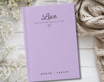 May include: A lavender hardcover journal featuring the black printed text Love WRITTEN ONE DAY AT A TIME with a small heart icon below it. The bottom of the cover displays the names alicia + trevor in black text on a smooth, solid surface.