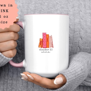May include: White ceramic mug with a pink interior and handle. The mug features a stack of illustrated books in shades of pink and orange, with the text "chapter 30 emma" below. The mug is 15 oz in size.