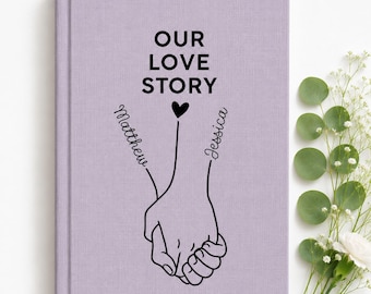 May include: A light purple hardcover book featuring the black text OUR LOVE STORY above a line art illustration of two hands holding each other. The names Matthew and Jessica are written in black script along the lines of the hands.
