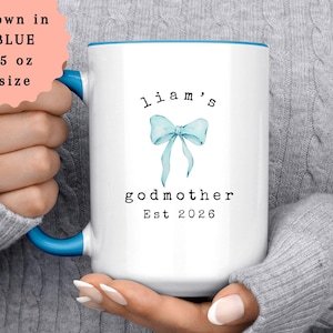 May include: White ceramic mug with a blue interior and handle. The mug features the text "Liam's godmother Est 2026" and a light blue bow illustration. The mug is shown in blue and is 15 oz in size.