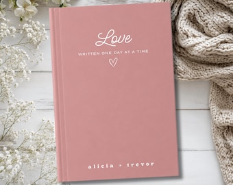 May include: A dusty rose colored hardcover book with white text that reads Love WRITTEN ONE DAY AT A TIME with a small heart outline below it and the names alicia + trevor printed in white at the bottom of the front cover.