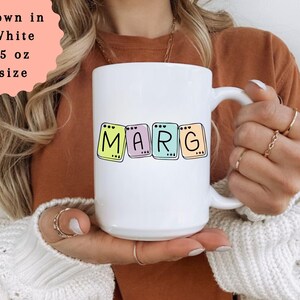 May include: White 15 oz mug with the name "MARG" in colorful, block letters. The letters are outlined in black and set against pastel backgrounds. The mug is held by a person wearing a brown shirt and a white sweater.