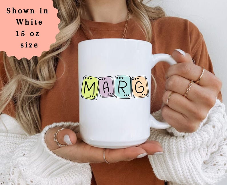 May include: White 15 oz mug with the name "MARG" in colorful, block letters. The letters are outlined in black and set against pastel backgrounds. The mug is held by a person wearing a brown shirt and a white sweater.
