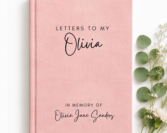 May include: A pink hardcover book with a fabric texture. The front cover features black text that reads LETTERS TO MY Olivia and IN MEMORY OF Olivia Jane Sanders. The book is positioned next to a sprig of green eucalyptus leaves and small white flowers.