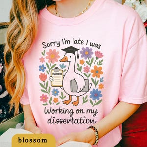 May include: Pink t-shirt with a graphic of a goose wearing a graduation cap, holding a clipboard and a book, surrounded by colorful flowers. Text reads "Sorry I'm late I was working on my dissertation."