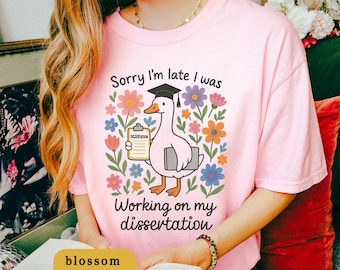May include: Pink t-shirt with a graphic of a goose wearing a graduation cap, holding a clipboard and a book, surrounded by colorful flowers. Text reads "Sorry I'm late I was working on my dissertation."