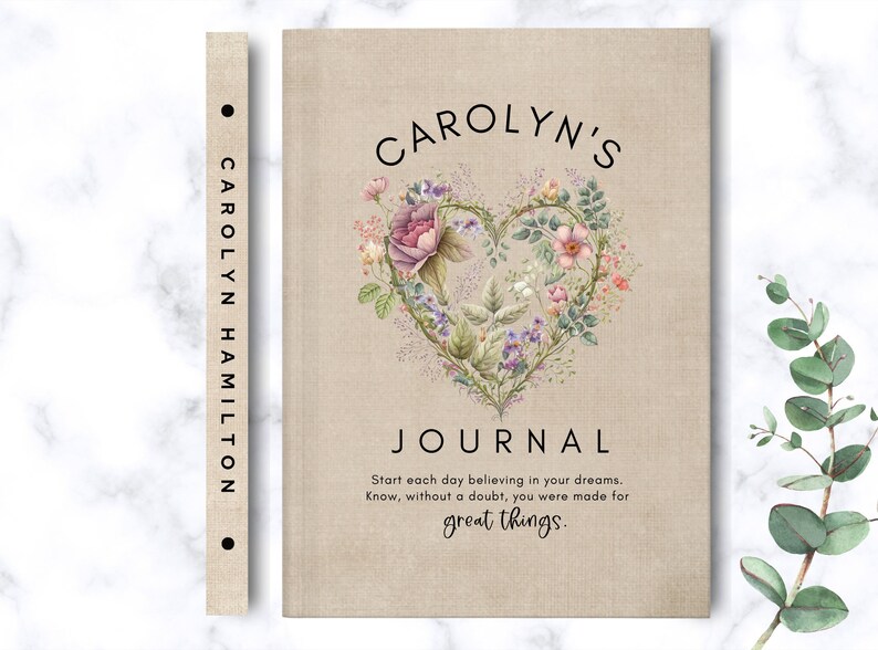 May include: A beige linen-textured journal featuring a heart-shaped floral illustration in pink, purple, and green. The cover displays the text CAROLYN'S JOURNAL and the quote Start each day believing in your dreams. Know, without a doubt, you were made for great things. The spine includes the name CAROLYN HAMILTON.