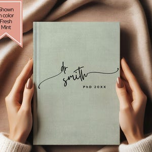 May include: A fresh mint-colored journal with the text "dr smith" in a cursive font and "PhD 20XX" printed below. The journal is held open by two hands.