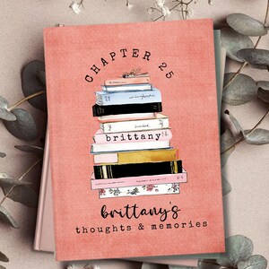 May include: A coral-colored journal with the text "Chapter 25" and "brittany's thoughts & memories." The cover features a stack of illustrated books in various colors. The journal is surrounded by greenery.