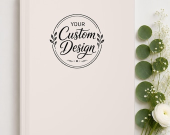 May include: A light beige hardcover notebook featuring a circular black graphic on the front cover. The graphic contains the text "YOUR Custom Design" in a stylized script font, framed by two small leaf sprigs on the left and right sides.
