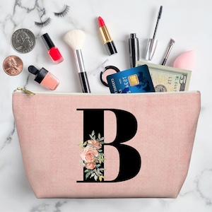 May include: A pink makeup bag with a large black letter "B" decorated with floral accents. The bag is filled with makeup, a credit card, and cash. Coins and makeup are scattered around the bag.