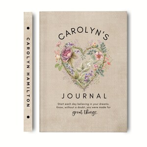 May include: A beige journal with a textured cover featuring a heart-shaped floral illustration, the text CAROLYN'S JOURNAL, and the quote Start each day believing in your dreams. Know, without a doubt, you were made for great things. The spine reads CAROLYN HAMILTON.