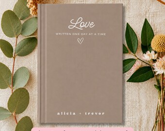 May include: A taupe-colored hardcover journal featuring the white printed text Love, WRITTEN ONE DAY AT A TIME, a small heart icon, and the names alicia + trevor at the bottom. The book has a vertical spine detail on the left side of the cover.