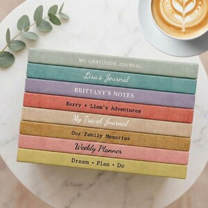 May include: A stack of eight hardcover journals in various muted colors including sage, teal, lavender, terracotta, beige, mustard, dusty rose, and olive. Each spine features unique white or black text including titles like My Gratitude Journal.