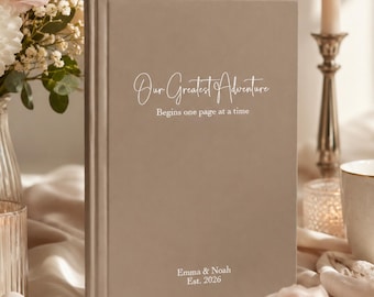 May include: A taupe-colored hardcover book featuring the white printed text Our Greatest Adventure Begins one page at a time and the names Emma & Noah Est. 2026. The book has a smooth, matte finish and a vertical spine detail on the left side.