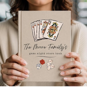 May include: A beige hardcover book titled The Moore Family's game night score book. The cover features a hand-drawn illustration of five playing cards showing an 8, A, J, K, and Q of hearts, along with a red die, a blue die, and two dominoes.
