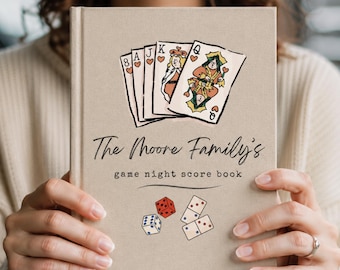 May include: A beige hardcover book titled The Moore Family's game night score book. The cover features a hand-drawn illustration of five playing cards showing an 8, A, J, K, and Q of hearts, along with a red die, a blue die, and two dominoes.