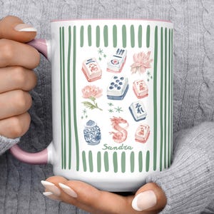 May include: A white ceramic mug with a pink handle and rim, featuring a green vertical striped border. The center design includes watercolor illustrations of mahjong tiles, pink flowers, a blue and white jar, a red dragon, and the name Sandra.