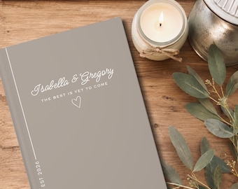 May include: A taupe-colored hardcover book featuring the white printed text Isabella & Gregory, the phrase THE BEST IS YET TO COME, a small heart outline, and the text EST 2026 positioned vertically along a thin white line on the left side.