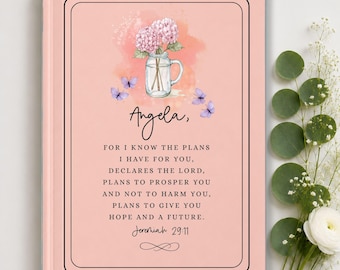 May include: A light pink hardcover journal featuring a watercolor illustration of pink hydrangea flowers in a glass mason jar with small purple butterflies. The text reads: Angela, FOR I KNOW THE PLANS I HAVE FOR YOU, DECLARES THE LORD, PLANS TO PROSPER YOU AND NOT TO HARM YOU, PLANS TO GIVE YOU HOPE AND A FUTURE. Jeremiah 29:11.