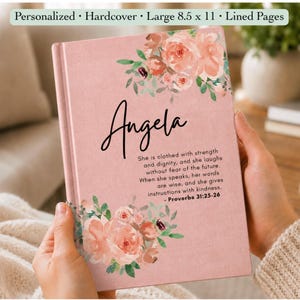 May include: A personalized hardcover journal with a dusty pink cover featuring watercolor floral illustrations in shades of peach and green. The name Angela is printed in black script, followed by the text of Proverbs 31:25-26 in black font.