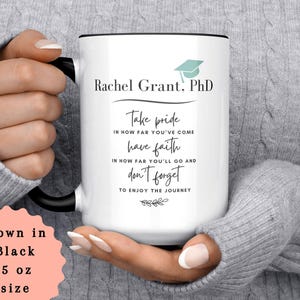 May include: White ceramic mug with a black handle and rim, featuring the text "Rachel Grant, PhD" and inspirational quotes. The mug also displays a graduation cap graphic. The mug is 15 oz in size.