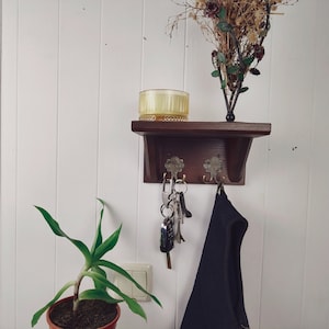 May include: A dark brown wooden wall shelf with two ornate hooks, holding keys and a black garment. A yellow glass container and a decorative floral arrangement sit on the shelf. A potted plant is in the foreground.
