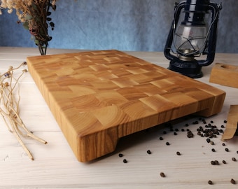 May include: A large, rectangular wooden chopping board with a chequered pattern. The board is made of light wood and has a natural finish. It is sitting on a white wooden surface.