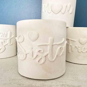 May include: Four white ceramic cylinder vases with embossed words and hearts. The vases read "Mom", "Sister", "The Best", and "Friends".
