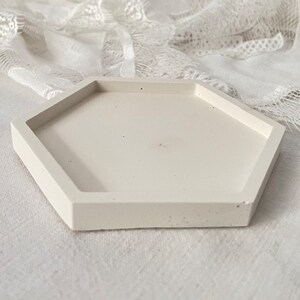 Peut inclure: Un plateau hexagonal en béton blanc avec une surface lisse légèrement texturée. Le plateau est parfait pour présenter de petits objets ou des bijoux.