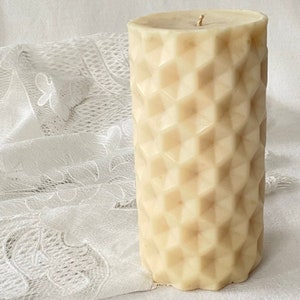 May include: A large, off-white pillar candle with a geometric, honeycomb-like pattern. The candle is sitting on a white lace tablecloth.