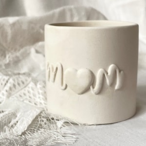 Puede incluir: Una taza de cerámica color crema con la palabra "Mom" en relieve en la parte delantera, con un corazón reemplazando la "o". La taza tiene forma cilíndrica y un acabado liso y mate. El fondo es blanco suave y borroso.