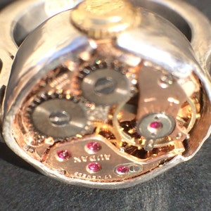 May include: A silver ring with a gold top and a clear glass dome revealing the inner workings of a watch. The watch face is visible through the glass dome and reads "17 JEWELS ADAN".