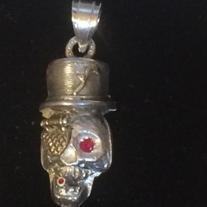 Pode incluir: Pingente de caveira de prata com um chapéu-coco e um olho de pedra preciosa vermelha. A caveira tem um design detalhado e um tapa-olho dourado.