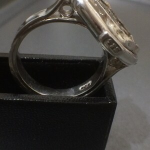 May include: A silver ring with a square-shaped setting featuring a detailed design. The ring is set in a black box.