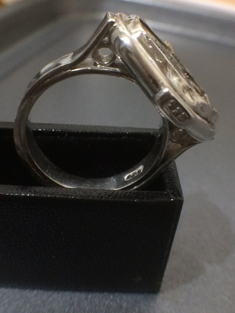 May include: A silver ring with a square-shaped setting featuring a detailed design. The ring is set in a black box.